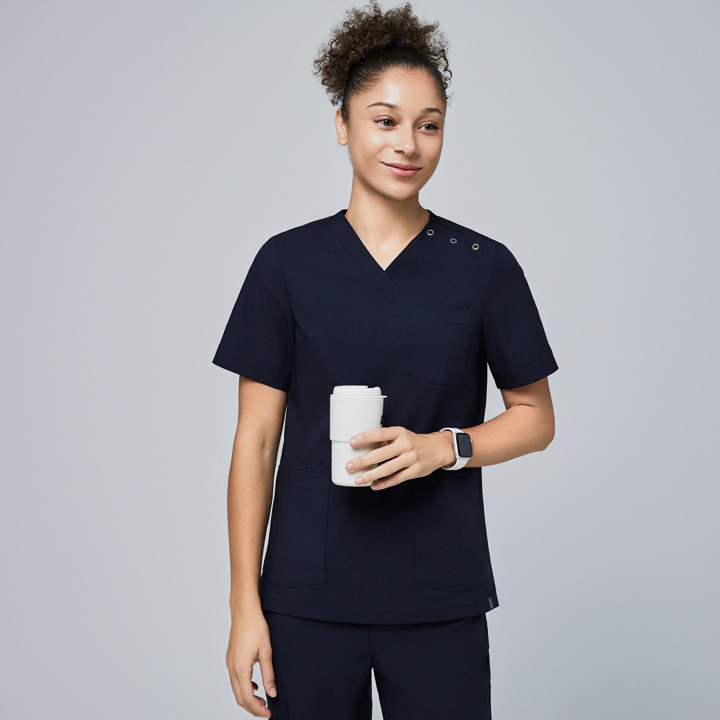 Image of Female model wearing a navy V-neck scrub top with two shoulder buttons and front pockets, holding a reusable coffee cup and smiling,Eco Navy